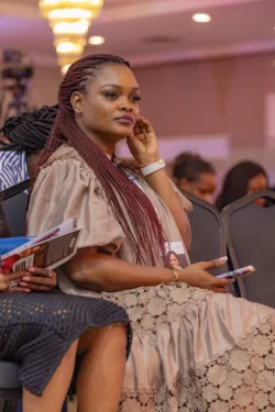 Seated woman with burgundy braids at a formal conference event, wearing a beige lace dress and badge. Formal event woman with burgundy braids, beige lace dress, and conference badge.